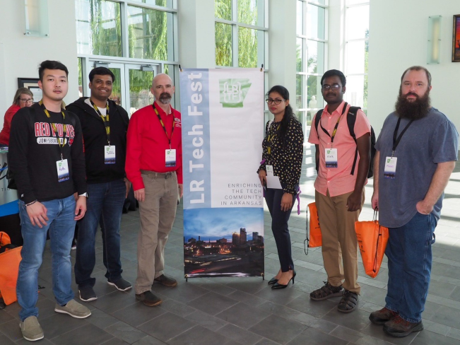 5 Computer Science students and a faculty member pose in front of the Little Rock Tech Fest Banner at the beginning of the event.
